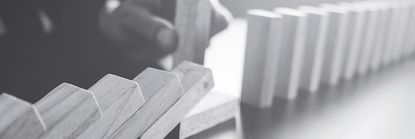 A line of rectangular wooden pieces stand up vertically on a table. Half of them have fallen over, like a line of dominoes. A hand holds one wooden piece in the background.