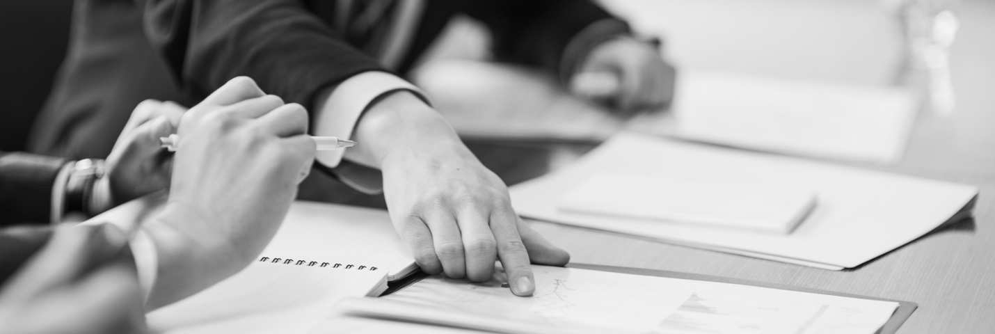 A man wearing a suit points to a chart on a piece of paper while sitting at a table with notebooks and pens.