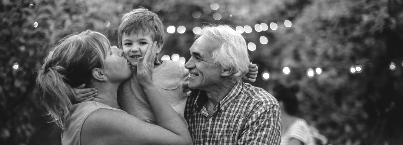 Grandparents hold their grandson. The grandma is kissing the grandson on the cheek.