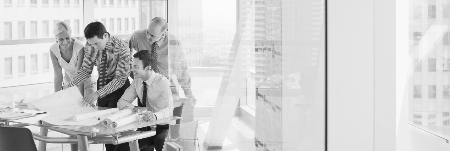 Four people look over a large sheet of paper as it is being unrolled across a modern desk. They are inside of an office with glass walls and windows. It is a very bright and minimal space.