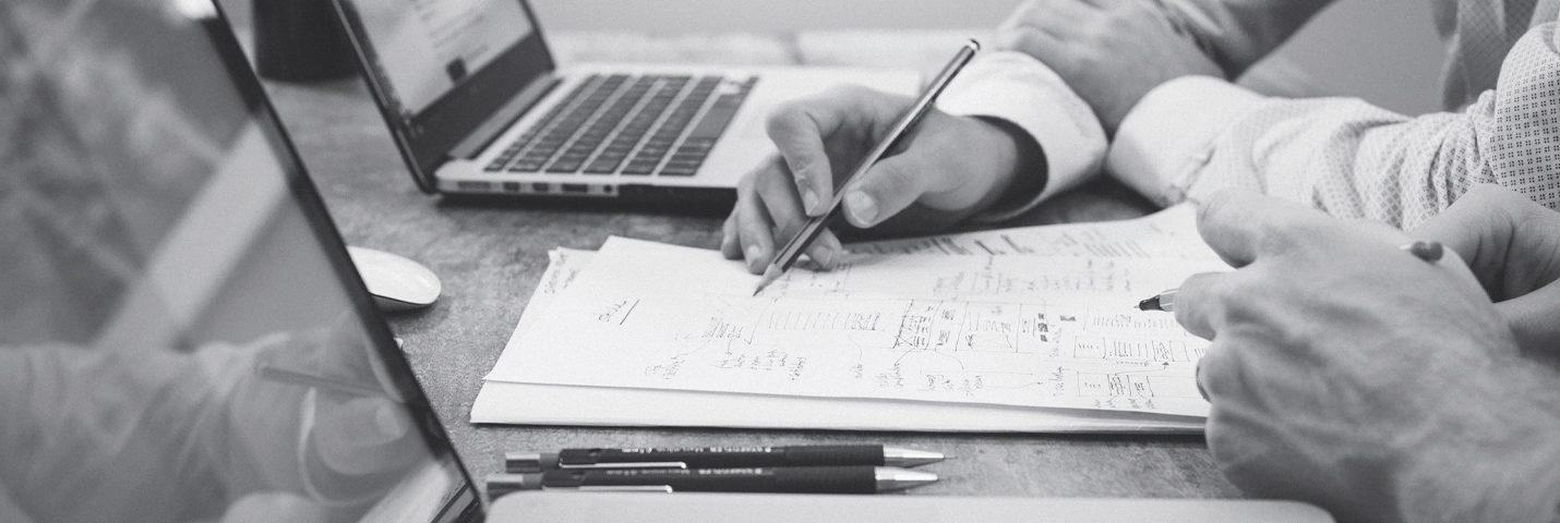 The hands of two people sitting at a table with their laptops, one holds a pencil and the other holds a pen, while pointing out various notes on a piece of paper.