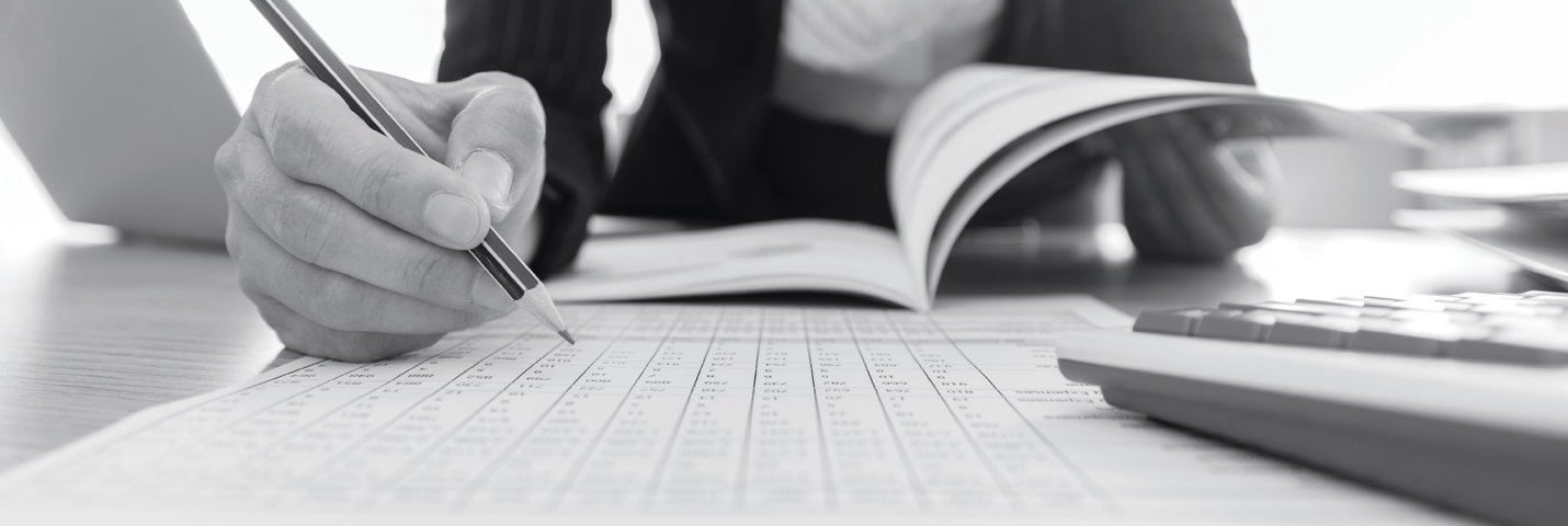 A spreadsheet sits on a desk, the top right corner partially covered by a computer keyboard. A person in a faintly striped blazer and white shirt leans over the desk with a pencil in one hand, hovering above the spreadsheet. The person flips through a thin booklet with the other hand.