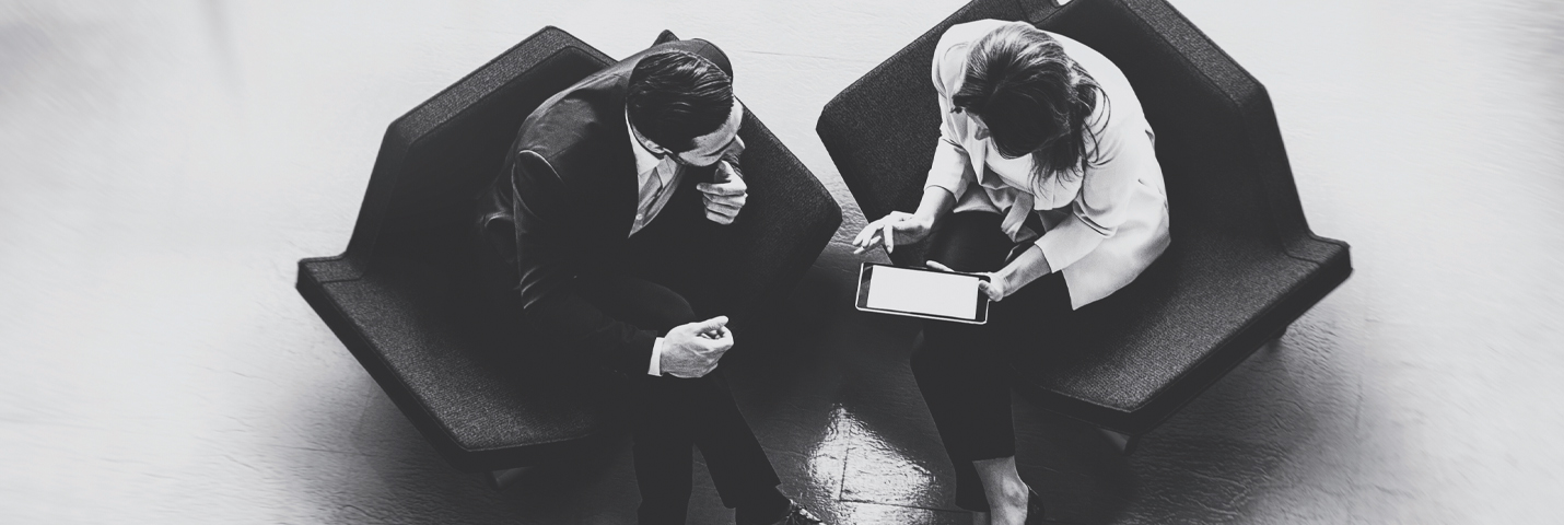 A man and a woman sit in comfortable unique geometric inspired chairs in an empty room as they look at a tablet the woman is holding. This image is taken from above, and looks down on top of them.