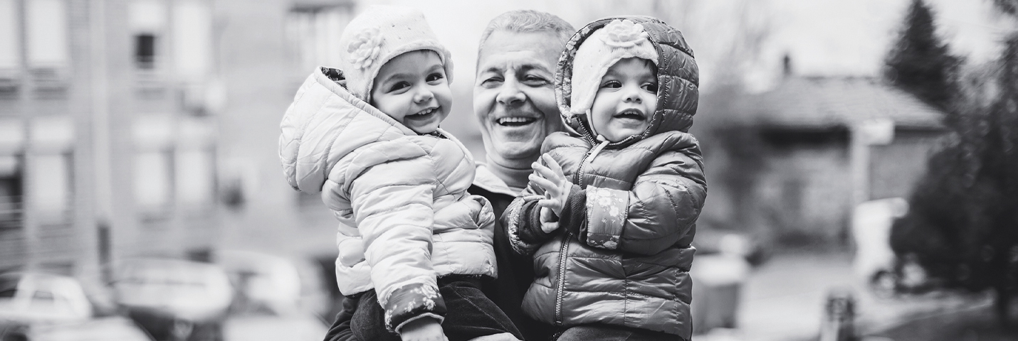 A man laughs while he holds twin toddlers who wear matching winter jackets and knit hats. The background is not in focus, but they are outside.